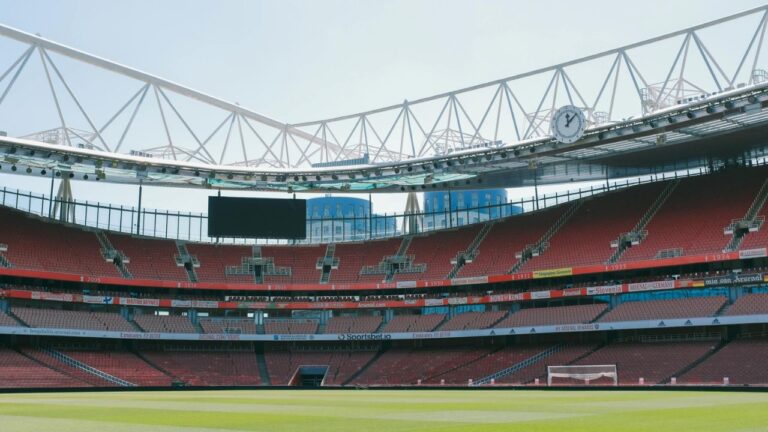Stort fodboldstadion med tribuner og grønt græs symboliserer både spillet på banen og det bagvedliggende økonomiske spil med massive investeringer