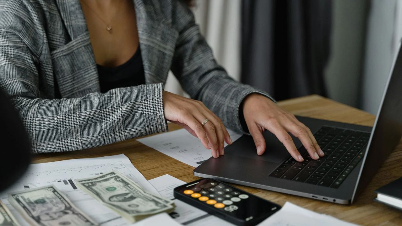 Kvinde sidder ved et bord med en Macbook, en iPhone, papirer med investeringsdata og pengesedler, der illustrerer kvinders effektivitet og engagement i investering