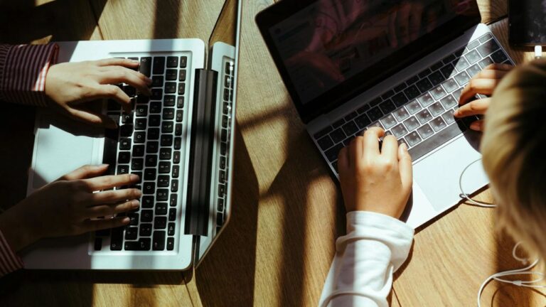 To unge piger sidder ved et bord med hver deres Macbook, hvilket symboliserer vigtigheden af at mestre økonomisk selvstændighed og investere tidligt i livet