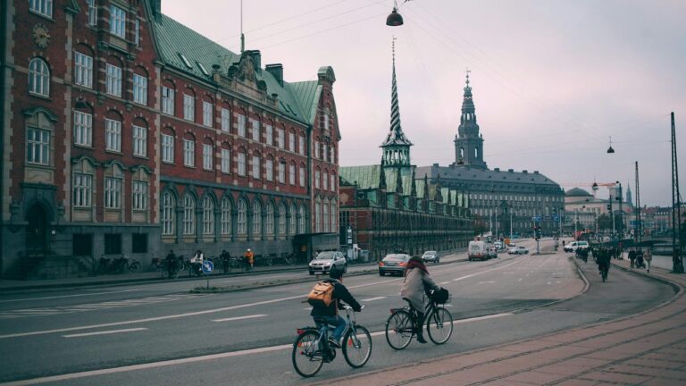 Cyklister og biler kører forbi den historiske bygning Børsen i København, hvilket symboliserer det danske C25-indeks, som indeholder de største danske virksomheder