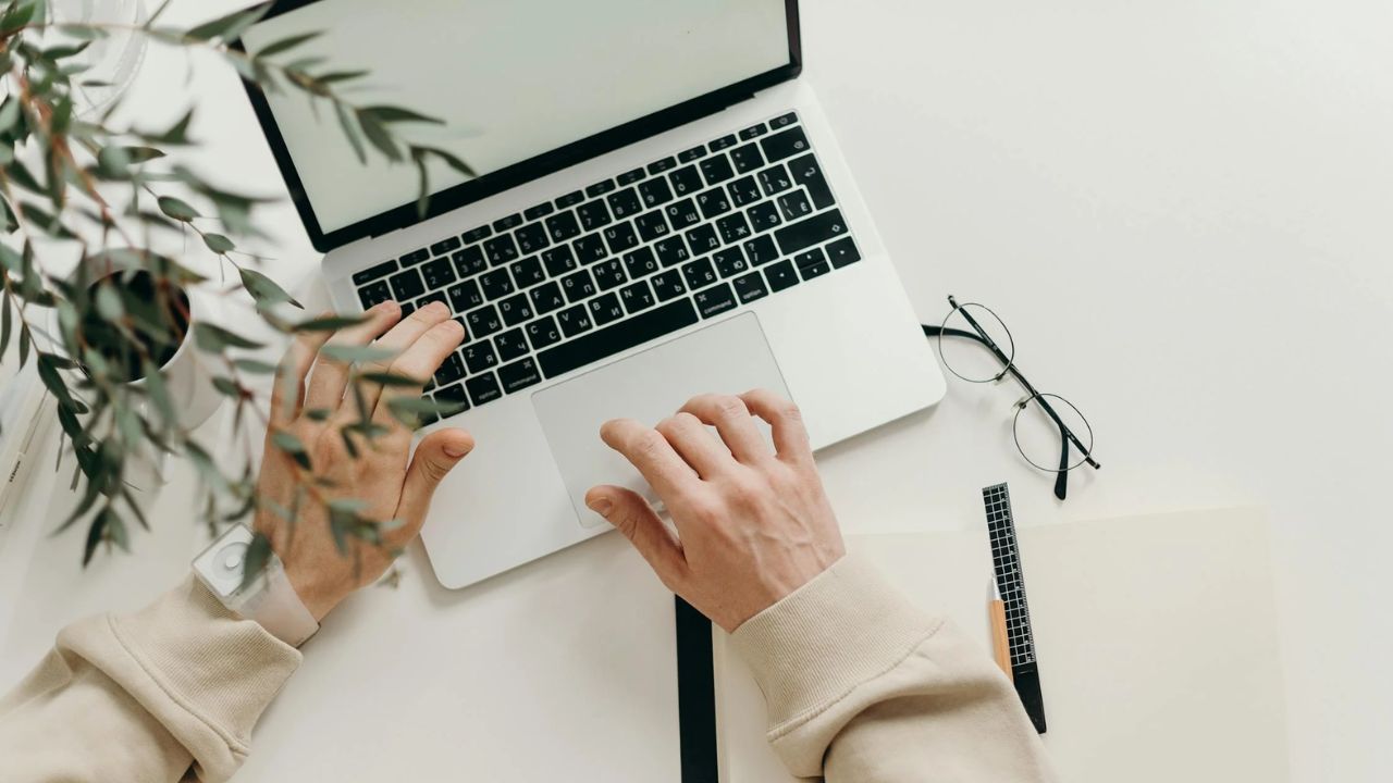 Person sidder ved et bord med en Macbook, en notesbog og et par briller, hvilket symboliserer arbejdet og forberedelsen, der kræves for at blive en god iværksætter