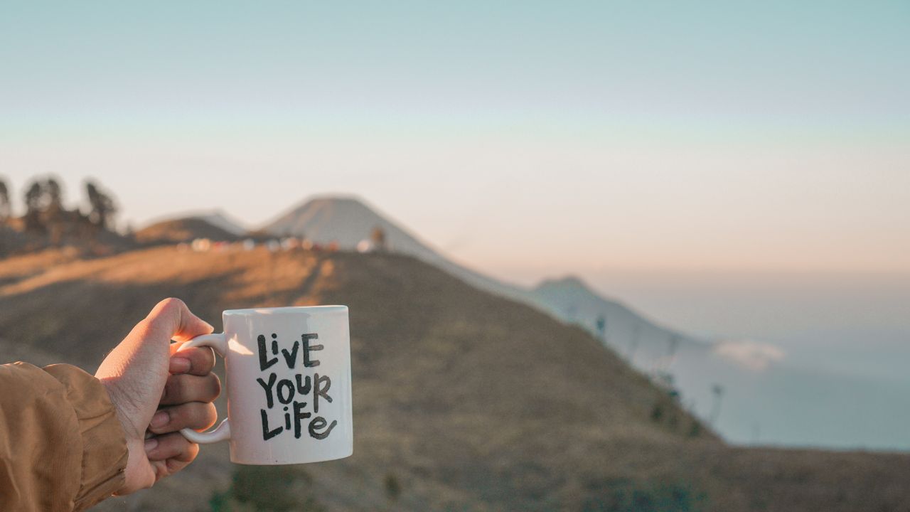 Person holder en hvid kaffekop med teksten 'Live Your Life' i et naturskønt område med bjerge, hvilket symboliserer drømmen om økonomisk frihed